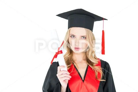 Serious young blonde woman in mortarboard holding diploma and looking at came Stock Photos