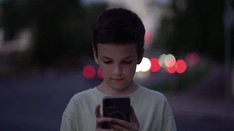 Serious young boy holding mobile phone with both hands on urban night street Stock Footage 340670642