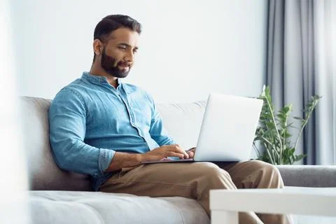Serious young eastern man working on laptop while sitting on sofa at home, hotel Stock Photos