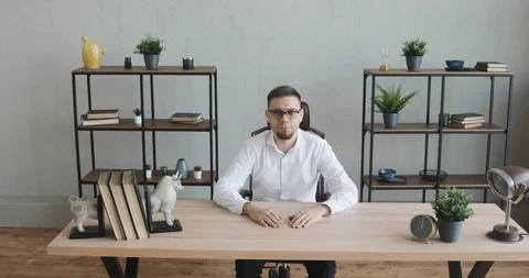 Serious young man office worker in glasses sitting at table looking at camera. Видео 120835754