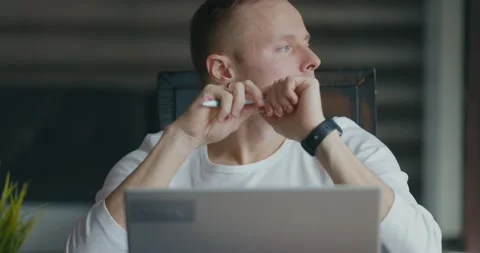 Serious young man sits at table and looks into laptop. Turns the handle in his Video stock 197576449