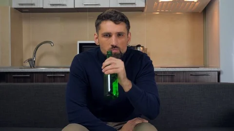 Serious young man sitting on the couch, drinking beer and watches TV Stock Footage 121840830