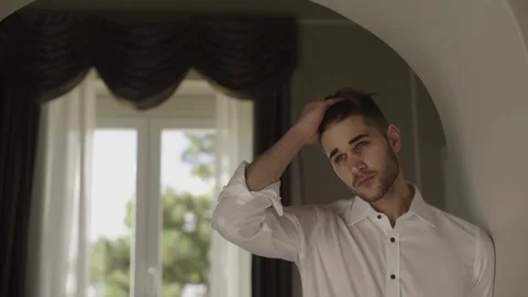Serious young man standing by the window and touching his hair Stock Footage 87146209