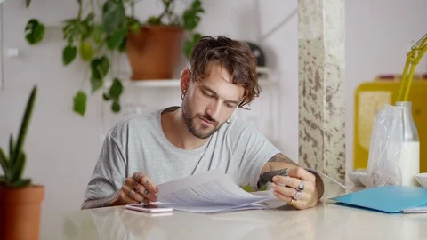 Serous pensive man sitting at kitchen and calculating invoices leafing papers at Stock Footage 108797679