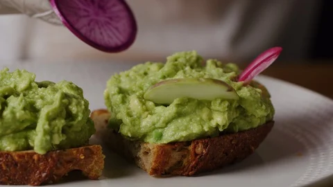 Serving avocado toast, putting slices of radish in squeezed avocado macro shot Stock Footage 128483370