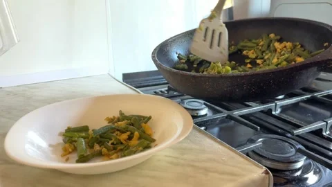 Serving cooked vegetables from frying pan into empty plate on kitchen stove Video stock 336524055