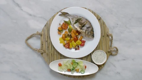 Serving roasted sea bream fish with vegetables, sauce, and fresh salad. Top view Stock Footage 122235539