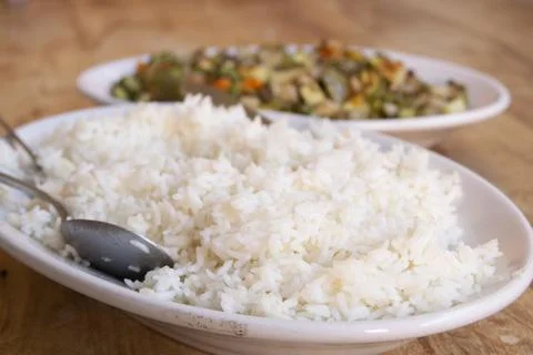 Serving spoons resting on fluffy white rice next to cooked vegetables Stock Photos