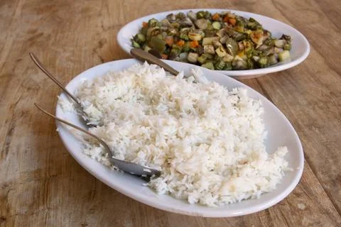 Serving spoons resting on fluffy white rice next to cooked vegetables Stock Photos