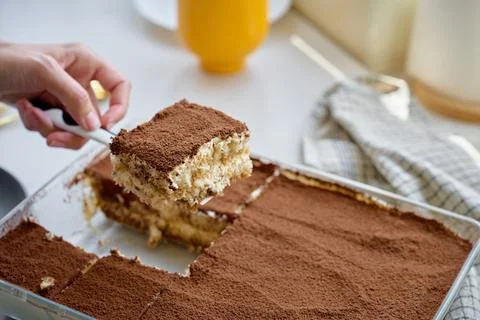 Serving tiramisu slice from a glass dish, lifted with a spatula after cutting Fotos Stock
