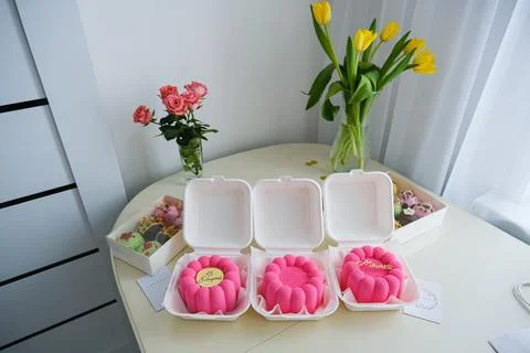 Set of three pink mini cakes in takeaway boxes on a table with flowers. Hom.. Stock Photos