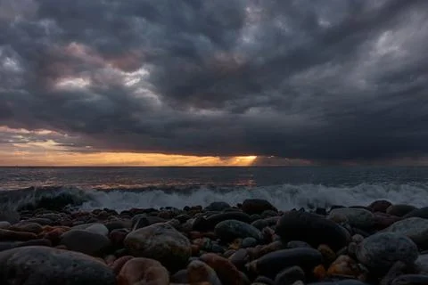 The setting sun breaks through low black storm clouds over the stormy sea. Stock Photos
