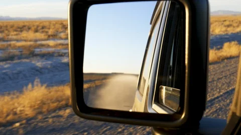 Setting sun illuminates clouds of dust behind moving vehicle in rear view mirror Stock Footage 120813105