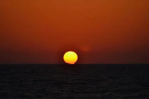 The setting sun touching a dark ocean against an orange sky. Stock Photos