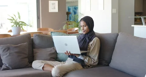 Settling on gray sofa, African American woman wearing dark hijab adjusting Stock Footage 341468476