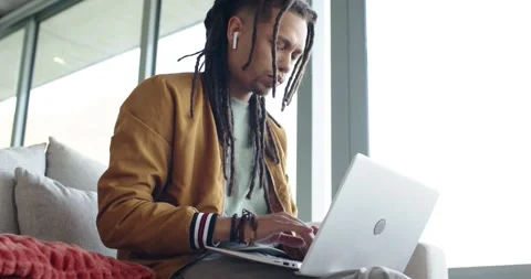 Settling on sofa by window, African American man in bomber jacket typing on Stock Footage 339161599