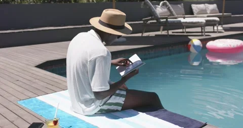 Settling on towel at backyard pool African American man relaxing, turning book Stock Footage 341736524