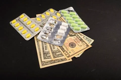 Several blisters with different pills and dollar bills on a black background. Stock Photos