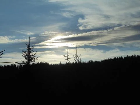 Several clouds on blue evening sky, covering the sun setting behind dark forest Stock Photos