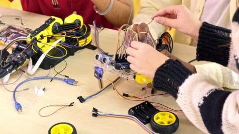 Several hands assemble a small wheeled robot on a table, connecting wires Stock Footage 325967433