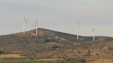 Several wind turbines rotating on a rocky, shrub-covered hill under soft Stock Footage 346703224