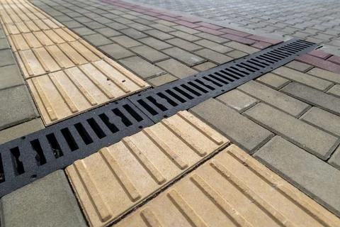 A sewer drain is located between two types of paving stones on a busy sidew.. 스톡 사진