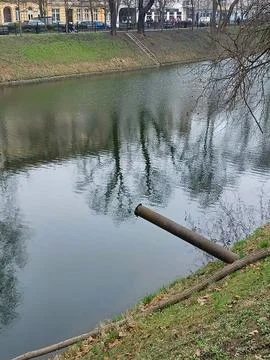 A sewer drain pipe hangs over a river in a city park. Environmental pollution Stock Photos