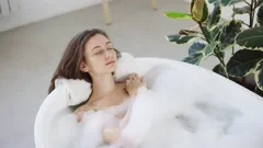 Beautiful girl in a bathtub with candles in glasses on the floor and fluffy  feathers on a white table. Stock Video Footage by ©petrunine #325087296