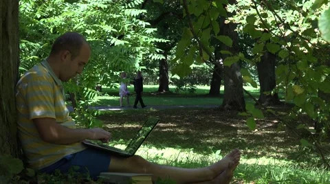 Shabby man working with laptop computer sitting under old tree in park. 4K Stock Footage 67752389