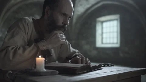 Shackled prisoner looking ahead thoughtfully from a book inside a gloomy cell Video stock 337648756