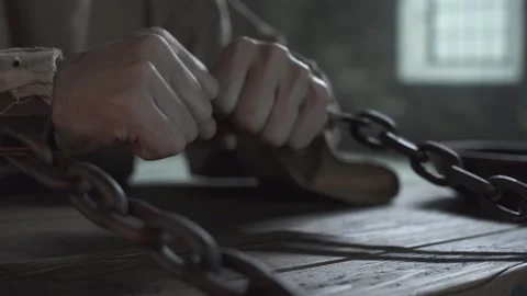 Shackled prisoner pulling iron chains in anger inside a gloomy jail cell Stock Footage 337648685