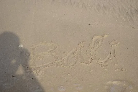 The shadow of a man on a light brown beach sand that says Bali. Stock Photos