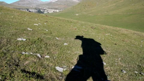 The shadow of a tourist walking uphill visible on a green meadow with rocks. Video stock 229277728