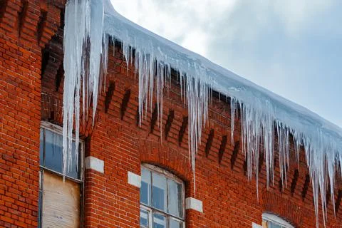 Sharp icicles hanging from snow-covered brick building roof, windows boarded Stock Photos
