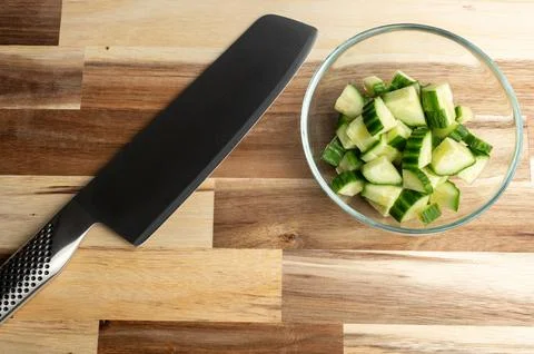 A sharp kitchen knife next to a bowl of chopped cucumbers on a wooden counter Stock Photos