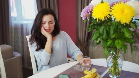 A sharp toothache from cold water in a brunette girl at the table at home. Stock Photos
