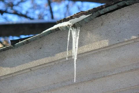 A sharp, translucent icicle hangs from a snowy roof edge against a textured w Stock Photos