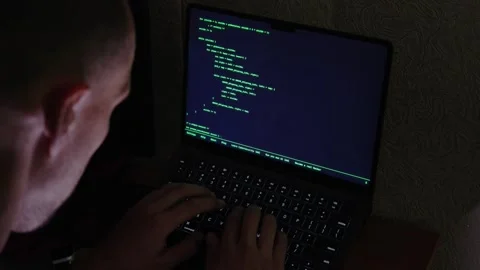 Shaved head man types code on laptop with green text in dim room Stock Footage 345521373