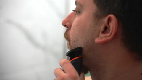 Shaving beard with electric trimmer, man in profile removing facial hair from Stock Footage 228683081