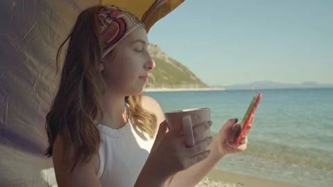 She spends time with her cell phone while drinking coffee in the camping tent. Vídeos de archivo 144323337