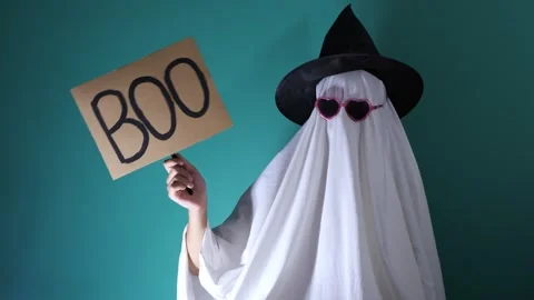 Sheet ghost in witch hat holding cardboard BOO sign on teal wall Stock Footage 343568512