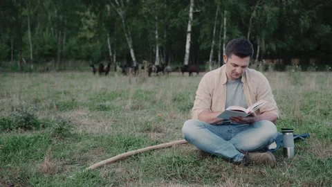 Shepherd sits on a grass reading a book against a herd of black sheep Stock Footage 235821165