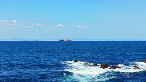 Ship sailing on the sea with waves and rocks visible at the shore on a clear day Stock Footage 344806982