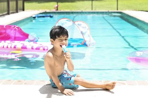 Shirtless boy eats popsicle while sitting poolside in summer sun Stock Photos