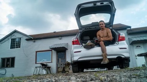 Shirtless Drone Pilot Sitting in Open Trunk of Hatchback Car Holding Remote Stock-Fotos
