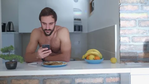 Shirtless man looking at his mobile phone in the kitchen 스톡 동영상 127648926