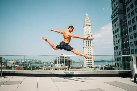 Shirtless man performs split leap on Boston rooftop with skyline view Stock Photos
