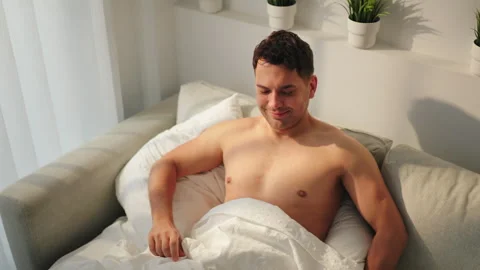 Shirtless man smiling in bed checks smartphone in sunny bedroom. Stock Footage 346800366