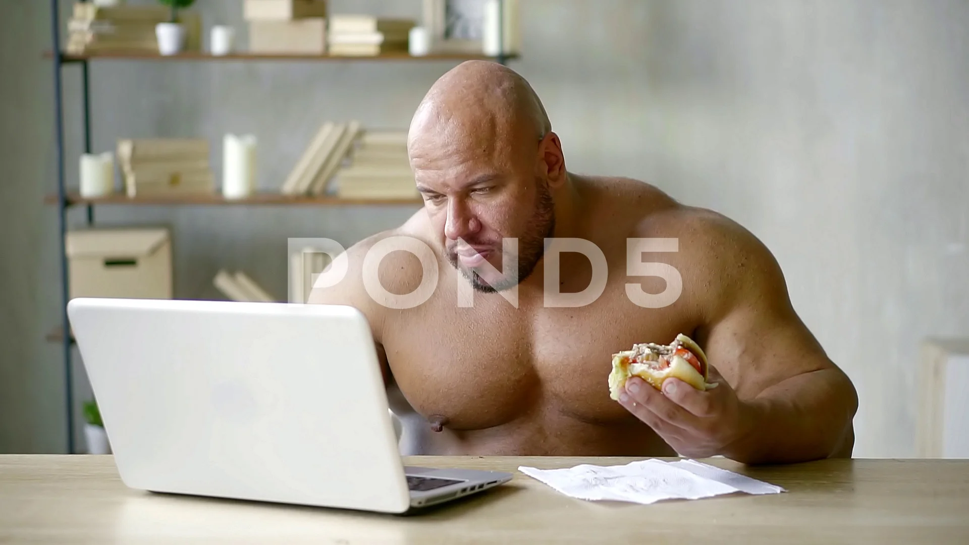 Shirtless weghtlifter holding fat junky hamburger while reading something  online