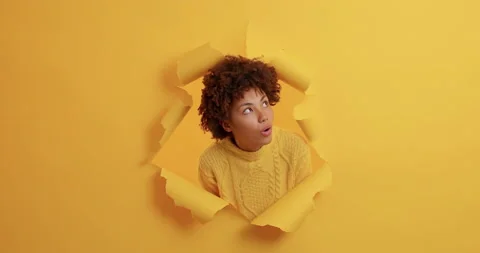 Shocked amazed dark skinned young woman appears in torn hole of paper background Stock Footage 143172344